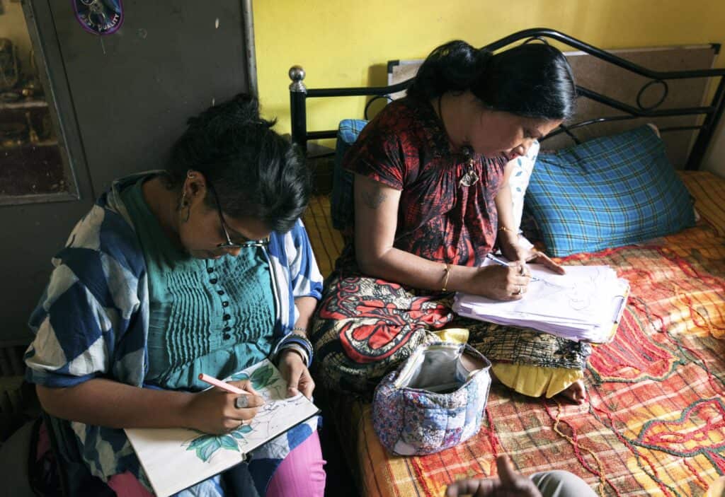 Two women sit next to each other while drawing. THey are wearing colorful garments and sitting in a colorful room||Noor has long dark brown hair and fair skin. She smiles at the camera in front of wooden doors and wears a neutral printed shirt.||||