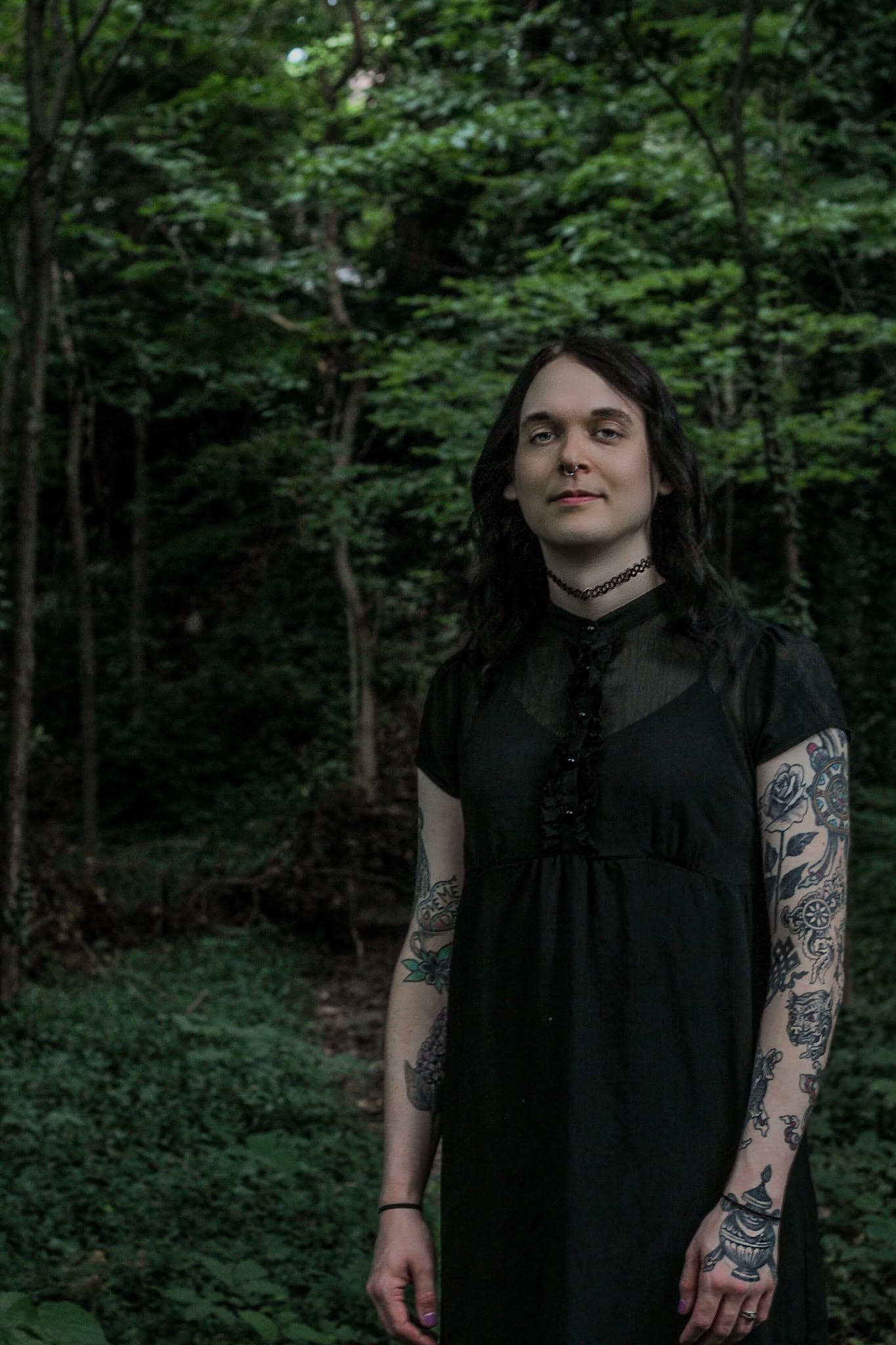 Grover wears a black dress and has fair skin. She has tattoos covering her arms and she stands against a lush green background. Her hair is black and flat. 