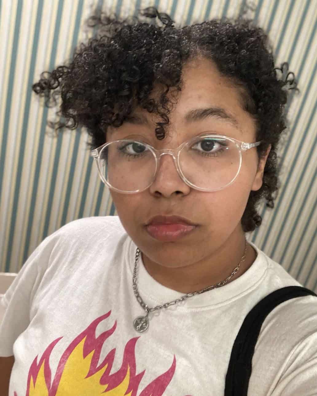 BMUD ANGEL in a selfie, they are wearing glasses and have their curly hair parted in a bang to the side with a stern look on their face