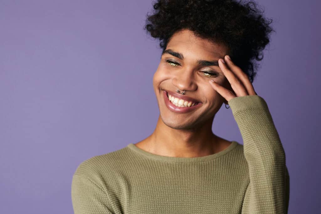 a genderqueer person with tan skins and curly black hair smiles in front of a purple background. They have glittery eye makeup.|