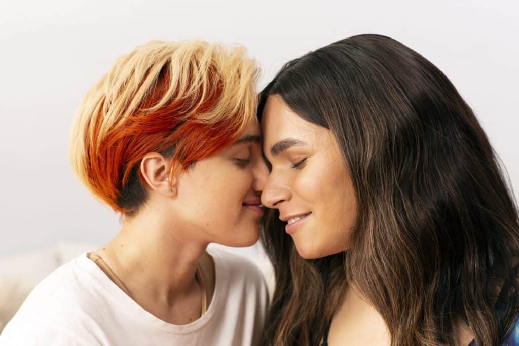 Two trans people lovingly resting their faces against each others. The fair skinned person on the left wears a white t-shirt and has short ombre blonde and red hair. The fair-skiiner person on the right has long brown hair and wears a black t-shirt.|