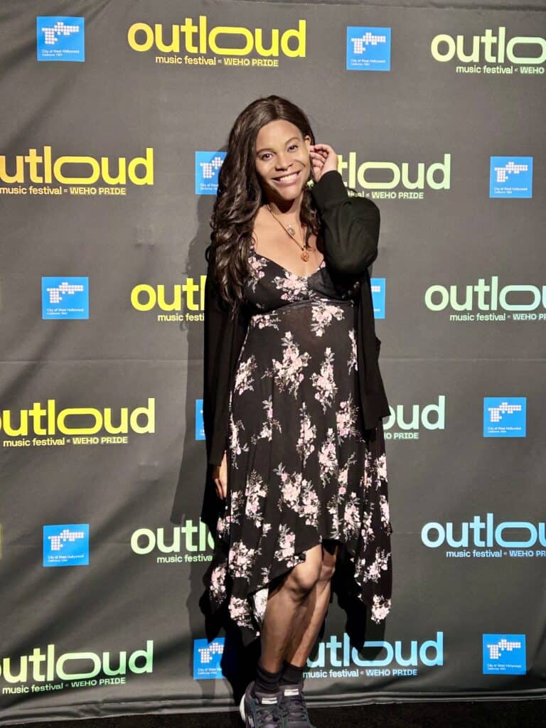 Blossom C. Brown in a black and pink dress posing and smiling on the step and repeat for the OUTLOAD musical festival. Credit: Blossom Brown