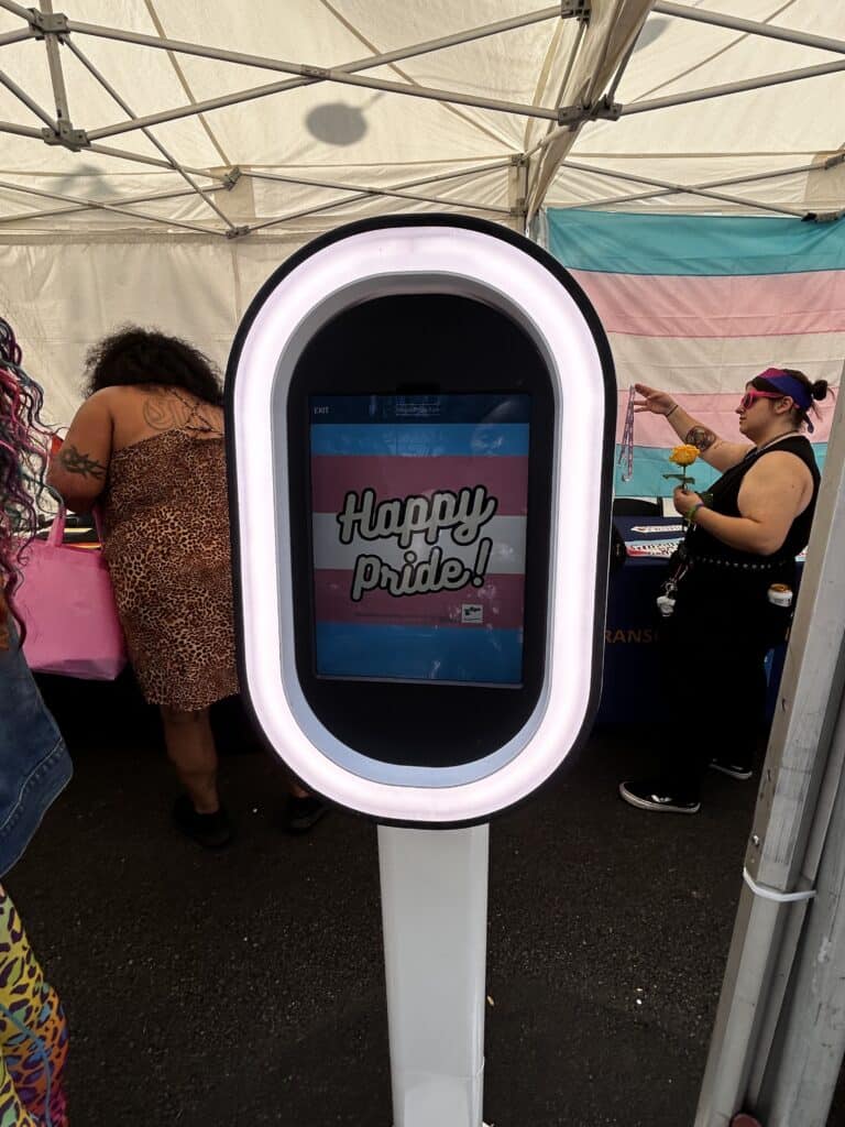 A photo of an virtual sign at a Street Fair booth reading “Happy Pride” in front of the trans flag. Credit: Blossom Brown