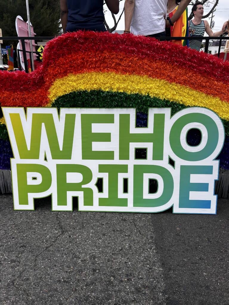 A photo of a cut-out green sign reading “WeHo Pride” on the Imperial Court of Los Angeles’ rainbow float at the Pride Parade. Credit: Blossom Brown
