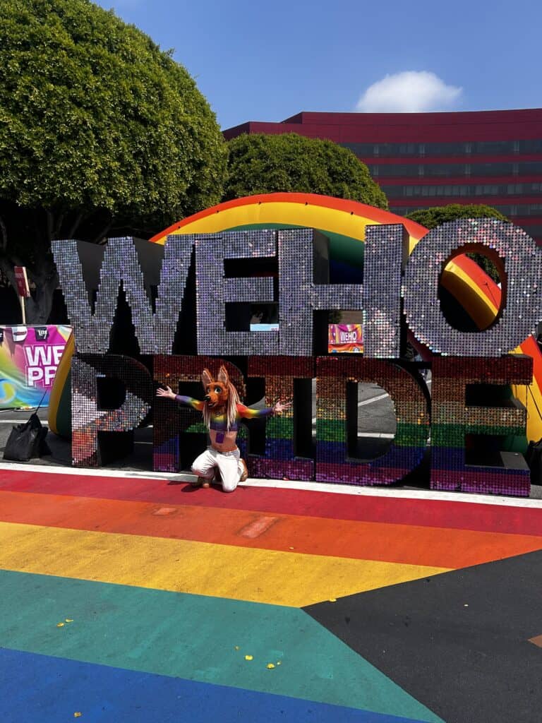 A person in a rainbow top wearing an animal mask poses in front of a large shiny, sequin sign reading WeHo Pride. Credit: Blossom Brown