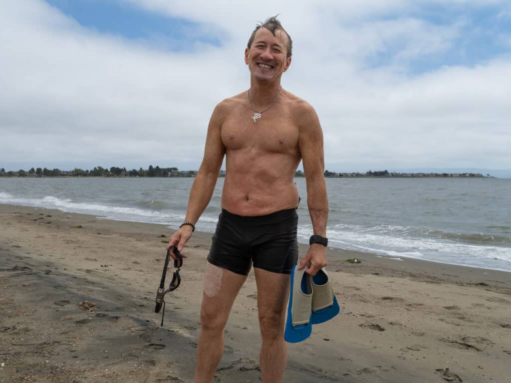 Willy Wilkinson standing on a beach holding a surfboard flippers.