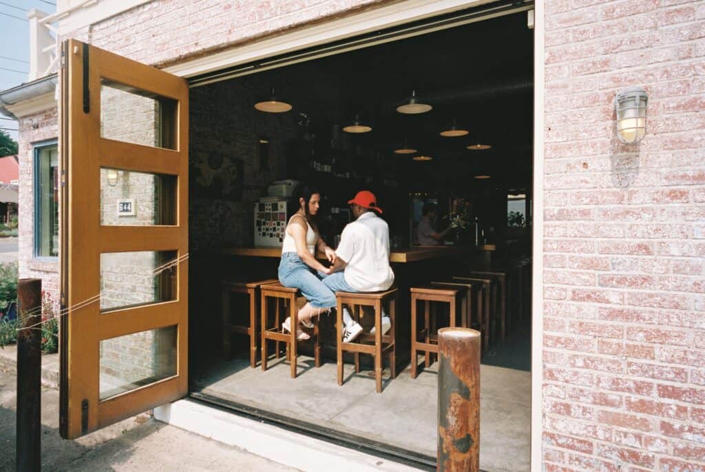 A wide shot ofMiss B Haven and Wilson inside a restaurant sitting on chairs and holding hands.