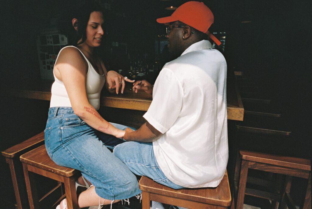 Miss B Haven and Wilson looking at each other and holding hands as they sit on chairs at the bar of a restaurant.