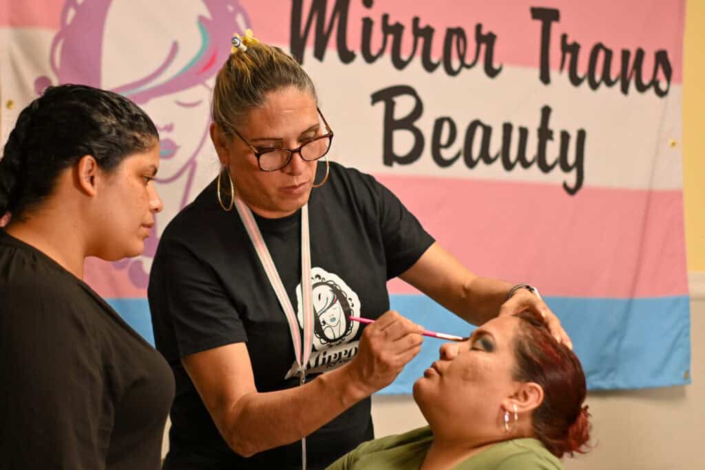A woman is getting her makeup done by Joselyn Mendoza in front of a transgender flag.