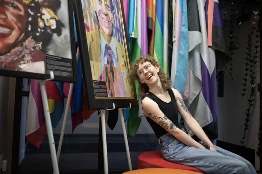 Morgan Peterson (they/them) takes a rest next to portraits of Marsha P. Johnson and Harvey Milk after setting up tables and chairs for an event at the Transformation Project office in Sioux Falls, SD.